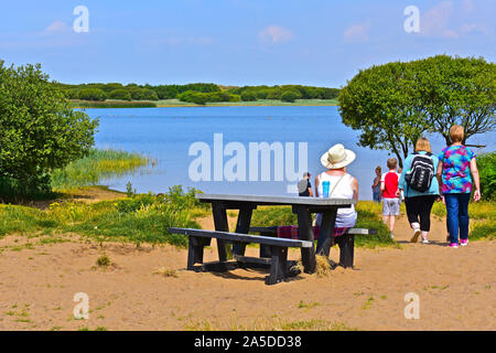 Une famille marcher dans le chemin de sable au bord de l'eau de piscine Kenfig, une réserve naturelle nationale site. Lady in white hat assis sur banc de pique-nique. Banque D'Images
