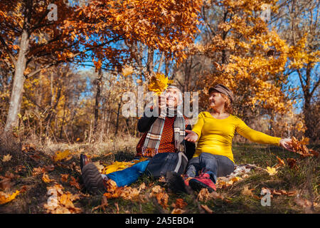 Saison d'automne de plaisir. Feuilles jeter Senior couple sitting in park. Heureux l'homme et de la femme de détente en plein air apprécier la nature Banque D'Images