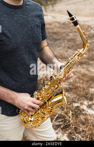 Jeune homme musicien joue du saxophone dans un lieu naturel au cours d'une journée à la campagne. Jouer instruments musique dans la nature sur un beau jour. Banque D'Images