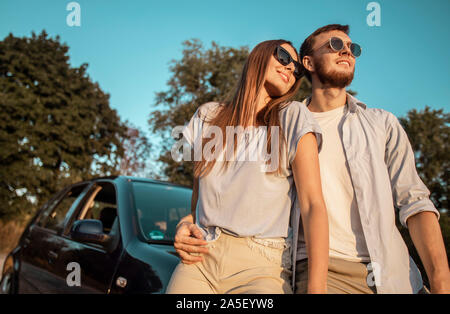 Couple aimant posant sur un coucher de soleil route de campagne appuyé contre un capot de voiture low angle view Banque D'Images