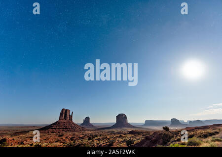 Vue grand angle célèbre buttes de Monument Valley au cours de crépuscule bleu nuit noire avec des étoiles dans la poussière de l'Arizona road et lune brillante dans le ciel Banque D'Images