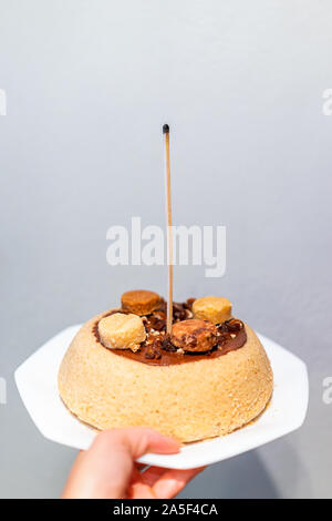 Main tenant une jaune vanille gâteau fait maison avec une sauce au chocolat et décoration bougie macaron avec match stick pour anniversaire Banque D'Images