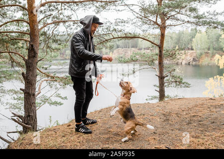 Jeune homme en blouson de cuir et jean noir donnant quelque chose de savoureux à mignon chiot beagle pure race, durant le frisson Banque D'Images