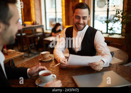 Portrait d'homme barbu du contrat de lecture et souriant joyeusement tout en parlant de partenaire au cours d'affaires au cafe Banque D'Images