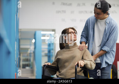 Portrait of young couple, femme en fauteuil roulant, à l'un l'autre et souriant tout en se tenant par les étagères de bibliothèque Banque D'Images