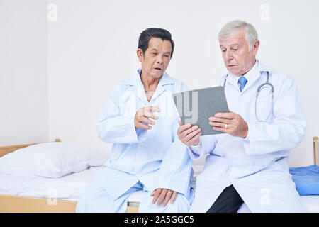 Doctor with tablet computer sitting on bed of senior patient et discuter avec lui des résultats de thérapie Banque D'Images