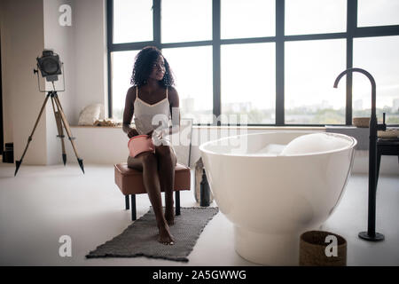 Curly excited woman sitting près de Bath avec mousse dans la salle de bains Banque D'Images