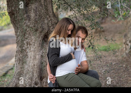 Pregnant couple assis à côté d'un arbre et à la recherche au ventre Banque D'Images