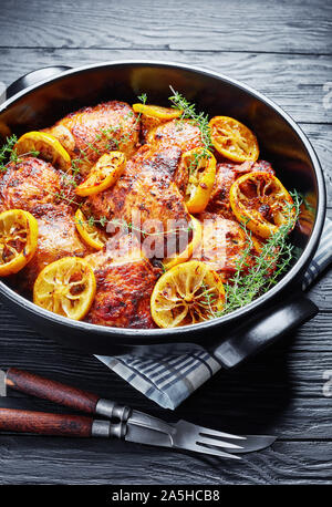 Cuisses de poulet frit avec des rondelles de citron et le thym dans une cocotte en céramique noire sur une table en bois, vue verticale Banque D'Images