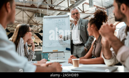 L'équipe d'affaires. Confiant businessman standing près de tableau blanc et d'expliquer quelque chose à ses jeunes collègues au cours de réunion au bureau Banque D'Images