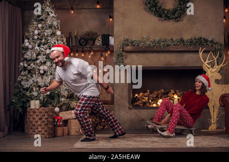 Image ofyoung man wearing Santa hat équitation femme sur un traîneau dans la chambre avec l'intérieur de Noël Banque D'Images