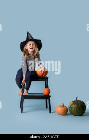 Fille en chapeau et des vêtements noirs se trouve sur escabeau avec citrouille à part Banque D'Images