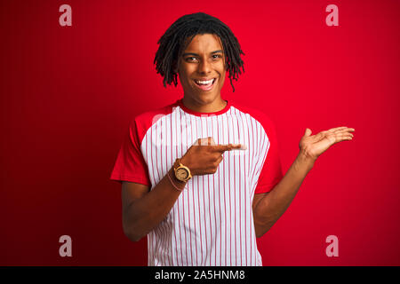 L'homme avec des dreadlocks Afro portant des T-shirt à rayures sur fond rouge isolé permanent étonné et souriant à la caméra tout en se présentant avec la main et Banque D'Images