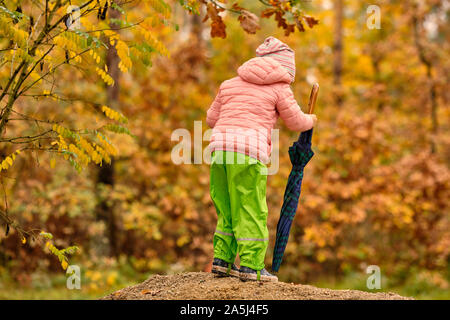 Vue arrière d'un enfant fille dans des vêtements chauds, des bottes de pluie et pantalon rubbert tenant un parapluie un jour de pluie en face d'une belle forêt d'automne en O Banque D'Images