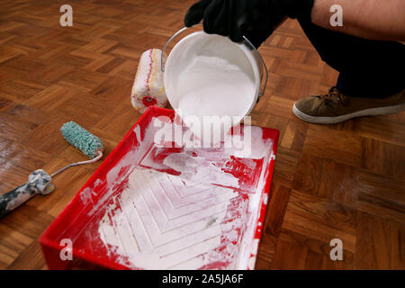 Travailleur de la construction de faire terminer la rénovation à l'appartement. Gants noirs avec peintre verse de couleur blanche de la benne dans le bol de mélange de rouge. Banque D'Images