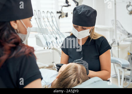 Vue du côté de dentistes femmes en uniforme et de masques dans les processus de guérison des dents. Petite fille couchée sur fauteuil dentaire alors qu'un médecin travaillant et l'examen de dent. Concept de traitement et de médicaments. Banque D'Images