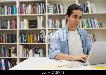 Portrait of young man using laptop latino-américains pendant leurs études au college library assis contre des étagères, copy space Banque D'Images
