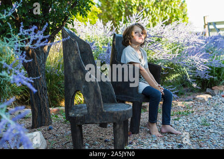 6 ans garçon assis dans un vieux fauteuil en bois dans le jardin formel Banque D'Images