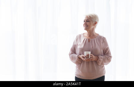 Vieille dame pensive avec mug d'un séjour près de window Banque D'Images