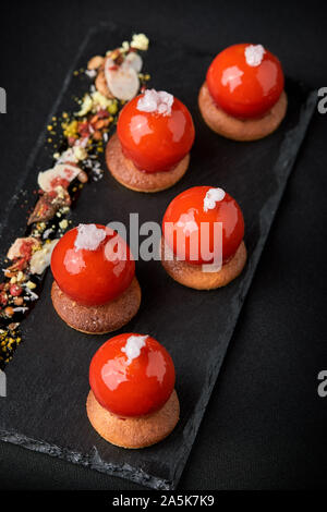 Mini tartelettes de fruits rouges sur le dessus vitré joliment disposés sur une plaque noire. Tartelette dessert festive moderne rouge et mousse dans le glaçage miroir shap Banque D'Images