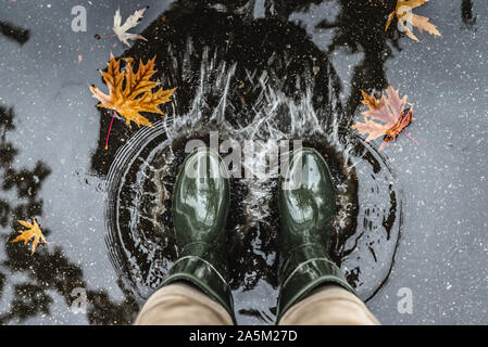 Les pieds dans des bottes en caoutchouc vert olive debout dans une flaque d'eau avec les feuilles tombées et de faire des éclaboussures. Banque D'Images