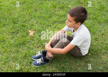 Vue latérale du garçon avec les cheveux bruns concentré bras croisés assis seul sur l'herbe. Enfant triste à la route. Les feuilles d'automne dans l'arrière-plan. Banque D'Images