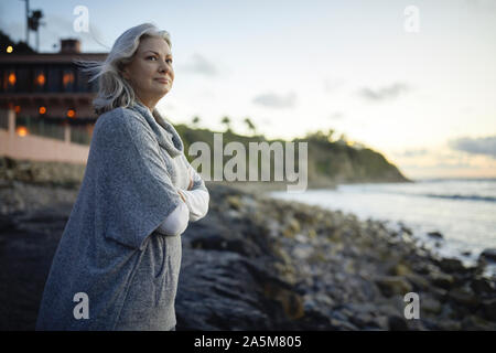 Vue latérale d'happy woman with arms crossed standing à Manhattan Beach contre le ciel au coucher du soleil Banque D'Images