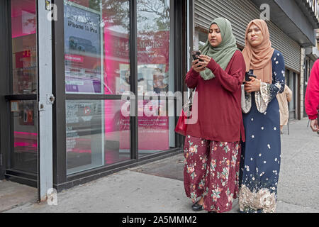 Deux jeunes femmes musulmanes portant le hijab et des vêtements traditionnels à pied sur la rue 74e tout en maintenant leur téléphone cellulaire. Dans la région de Jackson Heights, Queen, New York. Banque D'Images