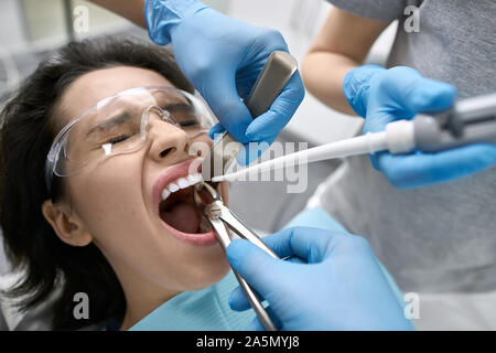 La souffrance girl aux yeux clos dans un bib du patient et des lunettes dans une clinique dentaire. Dentiste en bleu gants en latex est de retirer sa dent avec Banque D'Images