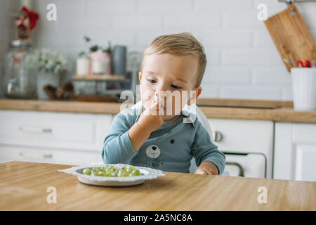 Charmant petit garçon peu concentré d'abord manger au raisin vert alimentaire cuisine lumineuse à la maison Banque D'Images