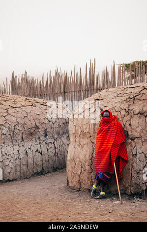 JUN 24, 2011 Serengeti, Tanzanie - Portrait de l'homme de la tribu Masai Masai ou en drap rouge Yeux regardant permanent de l'appareil photo par clay hut dans village. Banque D'Images