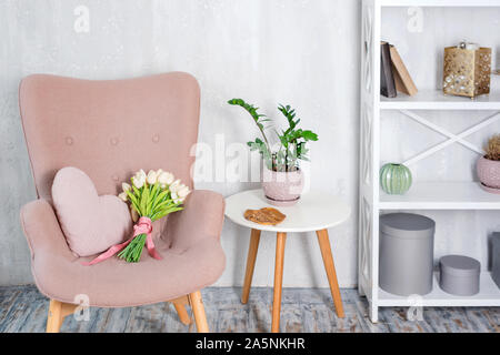 Rose élégant fauteuil avec coussin en forme de coeur dans un intérieur minimaliste. Décor en bois étagère avec près de mur gris. Bibliothèque avec des bougies Banque D'Images