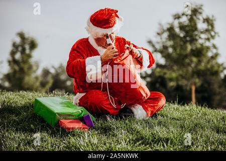 Stock photo verticale du Père Noël sans barbe accroupi sur le plancher l'organisation des cadeaux avec son sac rouge. Temps de Noël Banque D'Images