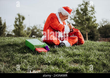 Stock photo verticale du Père Noël sans barbe accroupi sur le plancher l'organisation des cadeaux avec son sac rouge. Temps de Noël Banque D'Images