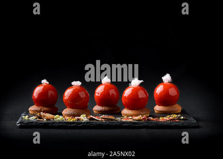 Mini tartelettes de fruits rouges sur le dessus vitré joliment disposés sur une plaque noire. Tartelette dessert festive moderne rouge et mousse dans le glaçage miroir shap Banque D'Images