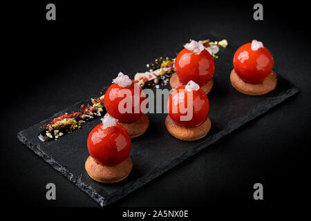 Mini tartelettes de fruits rouges sur le dessus vitré joliment disposés sur une plaque noire. Tartelette dessert festive moderne rouge et mousse dans le glaçage miroir shap Banque D'Images