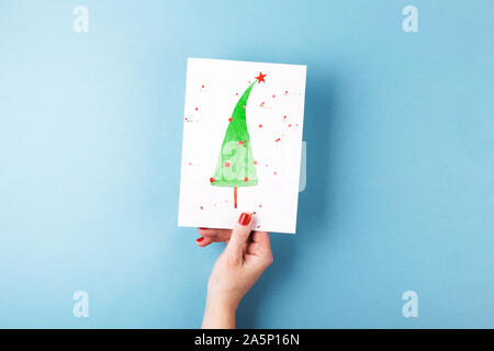 Vue aérienne sur femme's hands holding carte avec un arbre de Noël peints sur fond bleu. Composition de style minimal. Banque D'Images