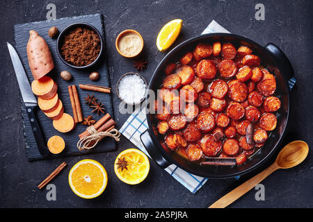 Confit d'ignames, patates douces cuites avec la cannelle, la cassonade et le beurre dans un plat de céramique noire. Ingrédients sur une pierre sur une table en béton, Banque D'Images