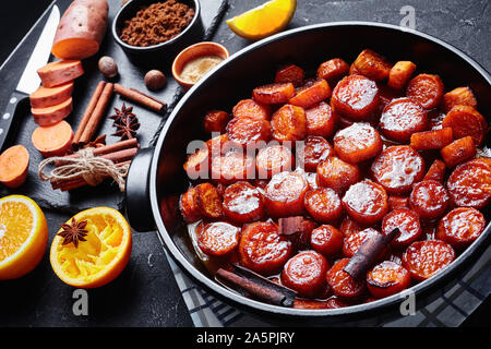 Confit d'ignames, patates douces cuites avec la cannelle, la cassonade et le beurre dans un plat de céramique noire. Ingrédients sur une liste de sélection sur une table en béton, Banque D'Images