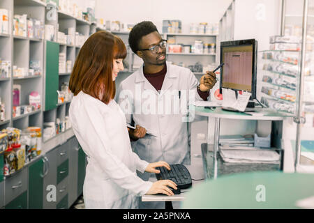Équipe de deux jeunes apothicaires en réputation debout devant des étagères avec les drogues et le travail sur ordinateur afin de faire des médicaments Banque D'Images