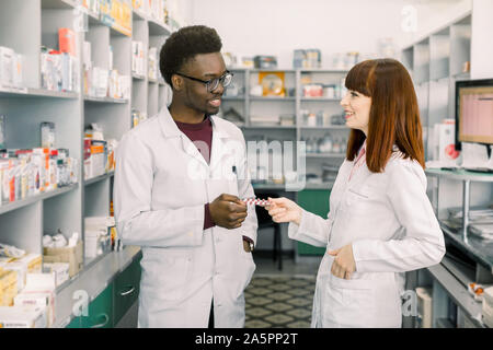 Certains pharmaciens masculins et féminins en pharmacie. Homme africain et Caucasian woman holding plaquette de pilules en position debout en pharmacie moderne Banque D'Images