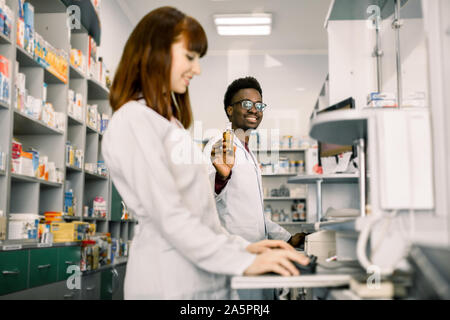 La médecine, pharmacie, soins de santé et personnes concept - heureux homme africain pharmacien montrant une bouteille de médicament à young Caucasian woman pharmacien à Banque D'Images