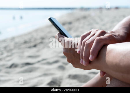 Libre d'un jeune homme de race blanche à l'aide de son smartphone sur une plage de sable blanc à côté de la mer Banque D'Images