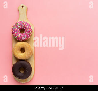 Trois différents beignets, beignets au chocolat, glaçage rose et sans glaçage sur une planche à découper en bois sur un fond rose Banque D'Images
