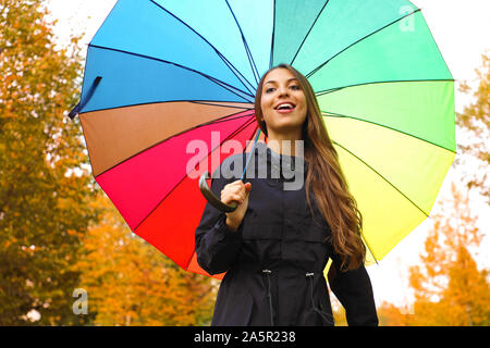 Jolie belle fille sous la pluie parapluie arc-en-ciel dans le parc. Young woman looking at camera avec parapluie coloré en plein air saison d'automne. Banque D'Images