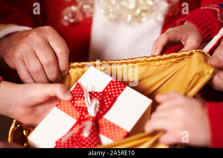 Le Père Noël ouvre un sac et donne des cadeaux aux enfants, assis près de l'arbre de Noël décoré avec des lumières - Joyeux Noël et bonnes fêtes ! Banque D'Images