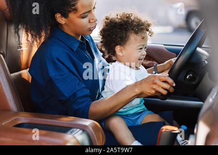 Tiré d'un adorable petit garçon assis sur les genoux de sa mère et maintenant le volant. Mère jouant avec son fils en voiture. Banque D'Images