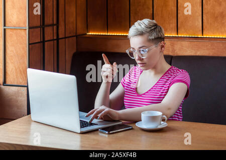 Portrait of serious young adult woman with blonde cheveux courts, en t-shirt rose et les lunettes est assis dans un café et montrant l'écran du doigt pour l'enseignement Banque D'Images