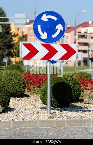 Bleu et blanc rond rond-point road signe au-dessus de rouge et blanc à rayures flèche tour dangereux panneau routier monté sur poteau de métal entourés de gravier Banque D'Images