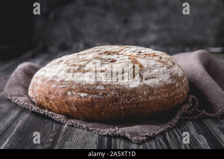 Un pain levain ronde avec le seigle et farine de blé. c'est posé sur une table en bois foncé, avec un fond sombre. Il y a un doux tissu en lin brun Banque D'Images